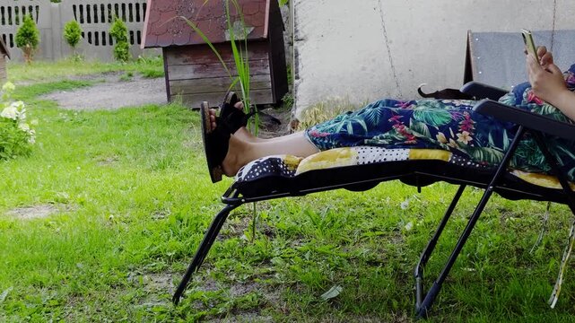 A woman in a floral dress resting on a reclining chair in a home garden, holding a smartphone, legs stretched out on a footrest wearing black sandals, green lawn, natural daylight.