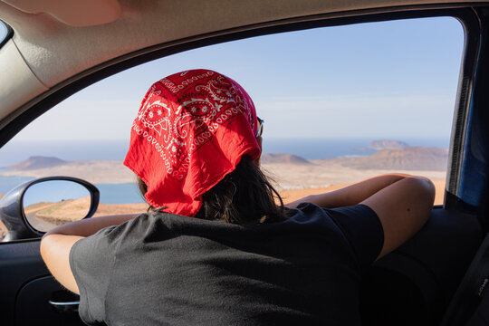 Traveler with red bandana leaning out car window, enjoying a panoramic island view and ocean during a summer road trip adventure