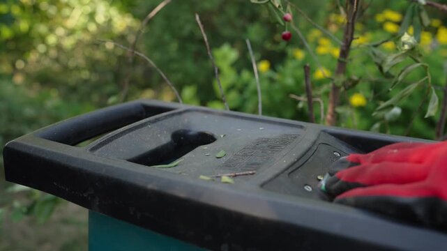 Garden shredder machine processing branches and leaves outdoors. Wood chipper hopper fed with plant material in a lush green garden setting.