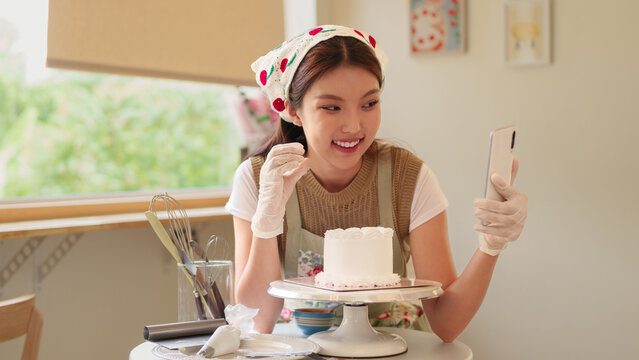 Young Asian baker filming smartphone video of decorated cake on turntable in bright home kitchen with copy space