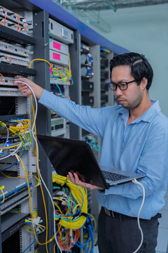 Focused Asian male IT network administrator uses a laptop to monitor server performance inside a bright data center managing complex corporate cybersecurity operations and infrastructure.