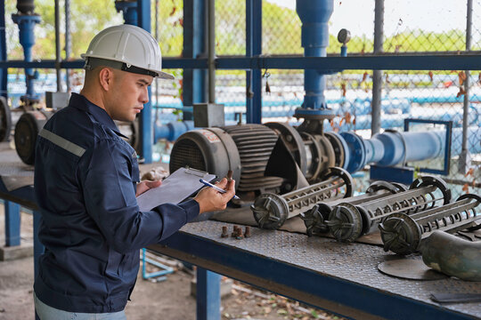 Focused male engineer wearing a hard hat writes on a clipboard while inspecting mechanical pump parts at an industrial water treatment plant ensuring equipment reliability and workplace safety.