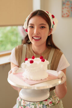 Young Asian woman proudly presenting homemade white frosted cake with cherry decoration on stand in bright kitchen