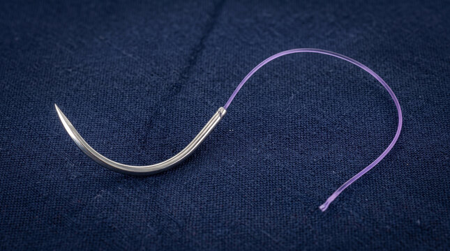 Stainless steel curved surgical needle with attached purple suture thread resting on a dark blue fabric