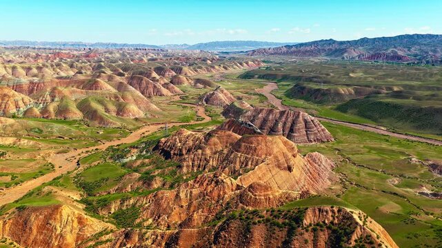 Aerial view of the dramatic Danxia landform with colorful striped mountains and green valleys under a blue sky in Xinjiang, China.