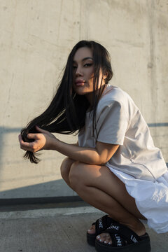 beautiful young Asian woman in a casual outfit and platform sandals crouches by a concrete wall, holding her long hair and looking confidently at the camera in warm sunset light.