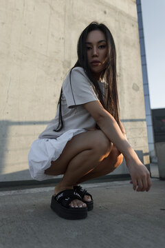 Low-angle portrait of a beautiful young Asian woman crouching on an urban rooftop in casual style clothing and black platform sandals, looking at the camera against a concrete wall.