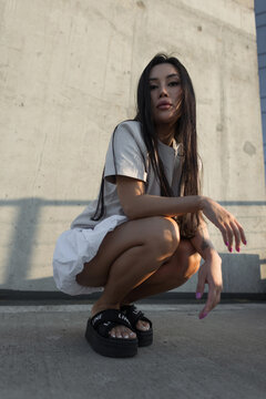 Confident young Asian woman crouching on an urban rooftop in trendy summer apparel and platform sandals. Low-angle shot during golden hour with a concrete background.