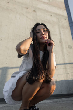 Low-angle portrait of a beautiful young Asian woman crouching on an urban rooftop, looking into the camera while touching her long hair during sunset against a textured concrete wall backdrop.