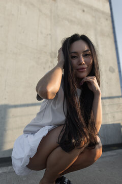Close-up portrait of a smiling young Asian woman sits on an urban rooftop, touching her long hair and chin in warm golden hour sunlight against a textured concrete wall.