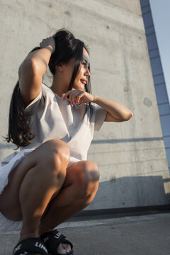young Asian woman in a casual beige t-shirt squats against a textured concrete wall, smiling and touching her long dark hair in warm afternoon sunlight. Perfect for lifestyle and urban fashion.
