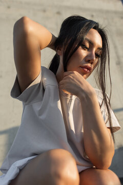 Close-up portrait of a pensive young Asian woman in a casual beige t-shirt, adjusting her long dark hair against a concrete wall in warm, dramatic afternoon sunlight. Ideal for urban lifestyle.