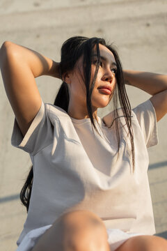 confident young Asian woman in a casual beige t-shirt holds her long dark hair back, looking away into the warm afternoon sunlight against a concrete wall. Perfect for urban lifestyle stock.