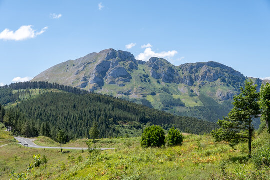A horizontal scenic view of the limestone peaks and lush green pine forests of the Basque mountains in Guipuzcoa, northern Spain, on a sunny summer day.