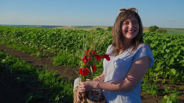 A happy young woman smiling while holding a beautiful bouquet of red poppies and wild grass in a lush green agricultural field during a sunny summer day