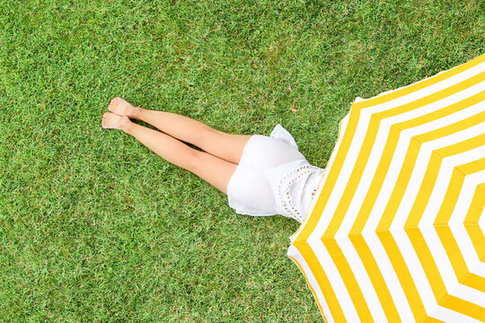 Woman in white dress lying under yellow umbrella on the green grass sunbathes at summer day. Top view, drone, aerial view.