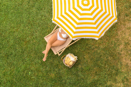Woman in a white bikini with basket for picnic sitting on deck chair under yellow umbrella  on the green grass sunbathes at summer day. Top view, drone, aerial view.