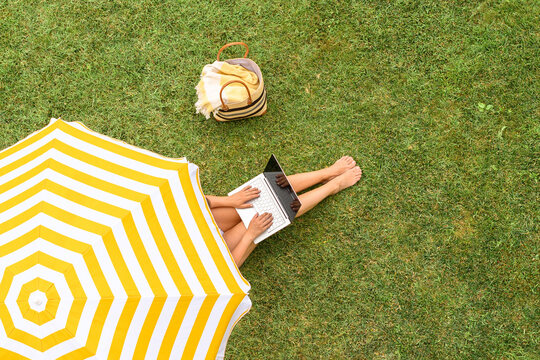 Woman in a white bikini with basket for picnic sitting on deck chair under yellow umbrella  on the green grass sunbathes at summer day. Top view, drone, aerial view.