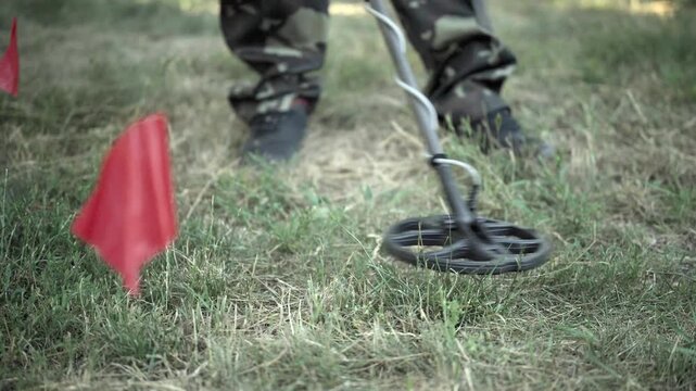A soldier in camouflage pants works with a metal detector near red flags on a grassy area, pointing out marked areas of interest for potential mine finds outdoors