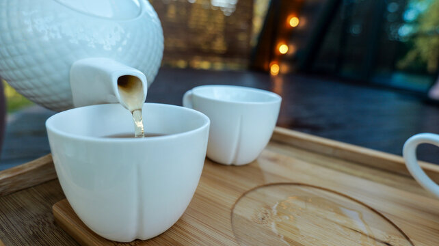 Pouring hot tea from a white teapot into a ceramic cup on a wooden tray