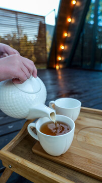 Pouring hot tea into a cup on a wooden tray with string lights in the background