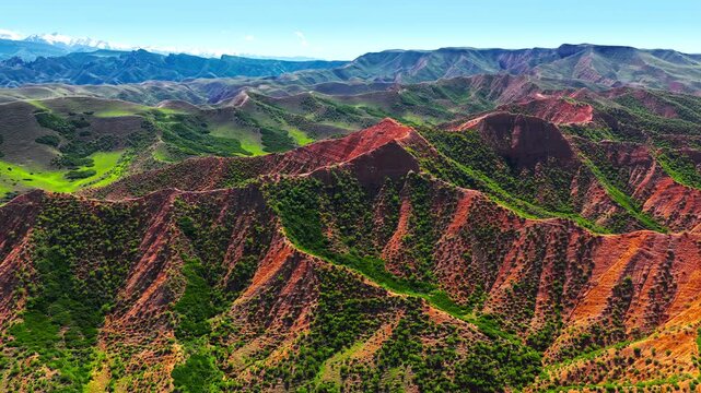 Aerial view of vibrant red sandstone formations and green valleys in Xinjiang, China, showing a dramatic and colorful mountainous landscape.