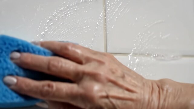 Close-up of a hand cleaning a tiled wall with a blue sponge.