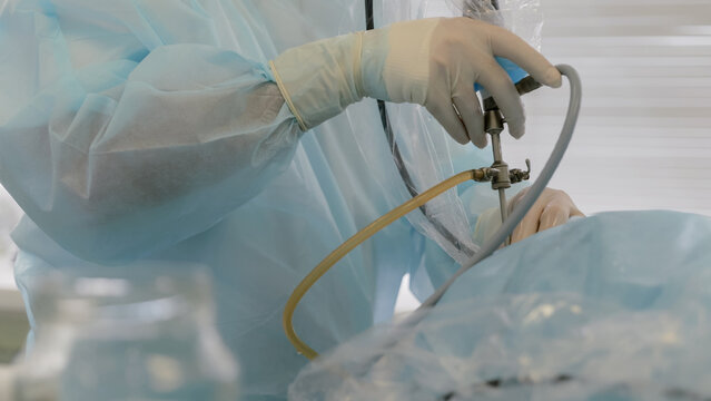 Gloved surgeon hands adjusting endoscopic instrument with clear tubing during minimally invasive procedure in sterile operating room with blue surgical drapes and bright clinical lighting