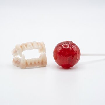 Halloween A pair of novelty vampire teeth rests beside a glossy red lollipop, hinting at a playful and perhaps spooky treat.