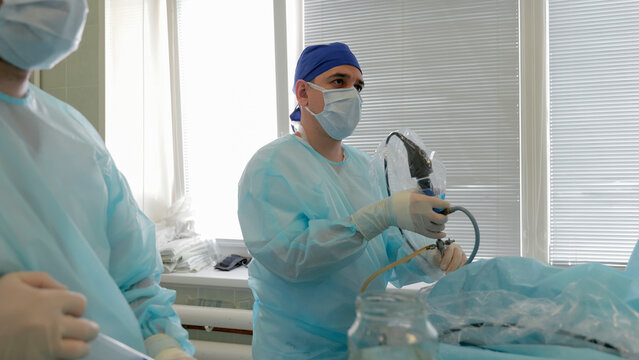 Surgeon and surgical assistant preparing endoscopic equipment beside draped patient in bright sterile operating room with medical trays, tubing, protective gowns, gloves, and focused clinical teamwork