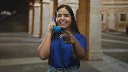 Fototapeta premium Young woman holds blue telephone to her ear and points finger directly toward camera in historic stone building; playful connection joyful.