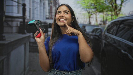 Woman holding a gamepad and points finger to cheek while smiling with controller raised in a street scene near parked cars and buildings  joyful play. © Krakenimages.com