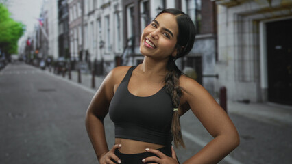 Fototapeta premium Sporty hispanic woman in black sports bra smiling with hands on hips on a city street; confidence vitality.