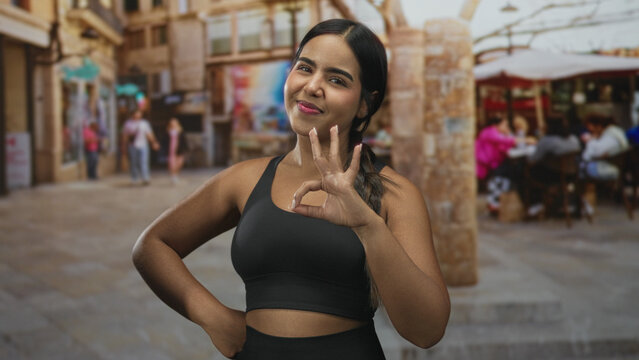 Young hispanic woman in black sports bra making ok hand sign with hand on hip on a street; fitness confidence.