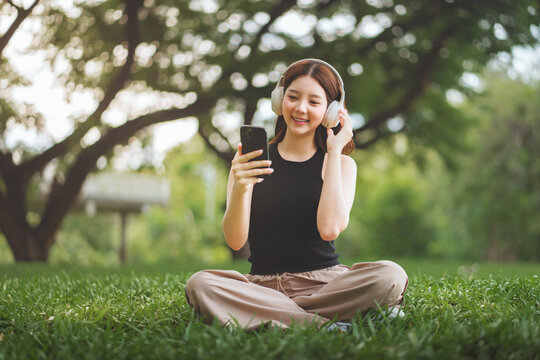 Cheerful asian woman wearing headphones and using smartphone to browse music or social media while sitting under a tree in city park during spring &ndash; relaxing lifestyle with soft sunlight, copy space