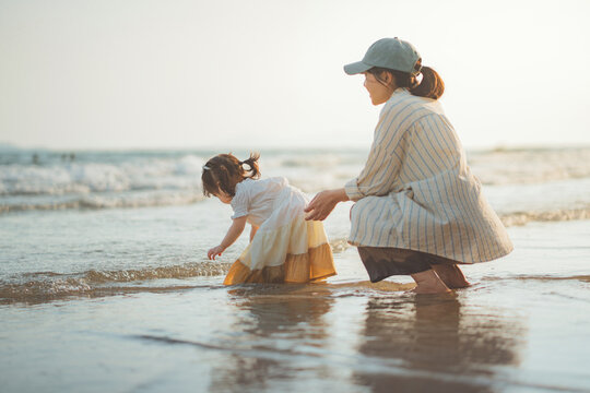 Side view of happy Asian mother watching her little daughter playing with sea waves on sandy beach, cute toddler child exploring water during family summer vacation for travel tourism, parenting.