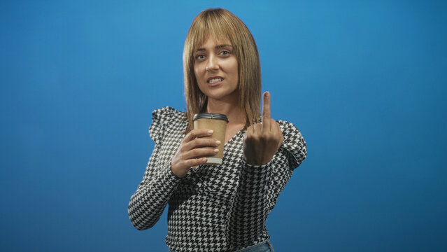 Woman holds a coffee cup and flips middle finger with hand in a blue studio, wearing houndstooth blouse and jeans, eyes half closed and smirking; sassy defiance.