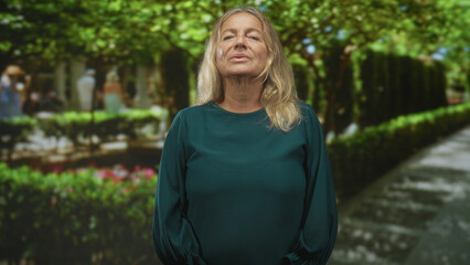 Fototapeta premium Senior woman face visible stands with chin raised in studio, hands relaxed at sides against leafy green garden path backdrop; confidence resilience.