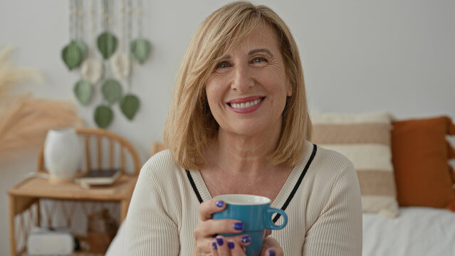 Woman middle age blonde holds blue mug with both hands, purple nails visible, smiling while seated on bed in a bedroom building; cozy serenity.