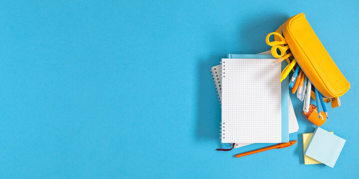 Open blank grid notebook with yellow pencil case and school stationery on blue background. Horizontal back to school banner template with wide copy space on left side, education concept background. 