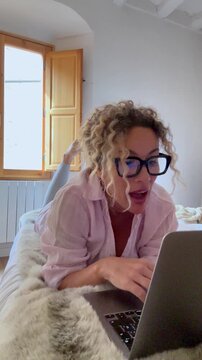 A young woman with curly hair and glasses lies comfortably on a bed, happily engaged with her laptop. She smiles and gestures, appearing to enjoy a video call or online interaction, highlighting remot