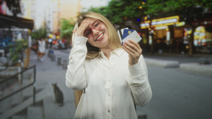 Fototapeta premium Woman holding a bunch of credit cards, hand to forehead gesture and smiling on a busy city street with shop lights and pavement; relief savings.