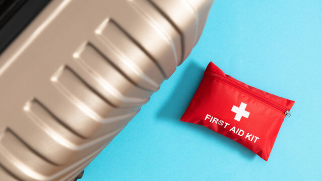 Red first aid kit pouch next to plastic travel suitcase on blue background. Medical travel insurance concept, safe trip banner with copy space, vacation health care, luggage preparation layout.