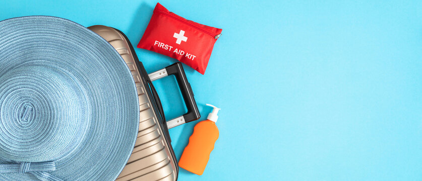 Red first aid kit, straw hat and sunscreen bottle on suitcase. Medical travel insurance concept, safe summer vacation banner with blue background and wide copy space for advertising text.