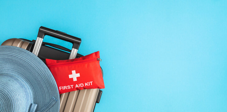 Red first aid kit pouch and straw hat on travel suitcase over blue background. Medical travel insurance concept, safe summer vacation banner with side copy space, medication packing backdrop.