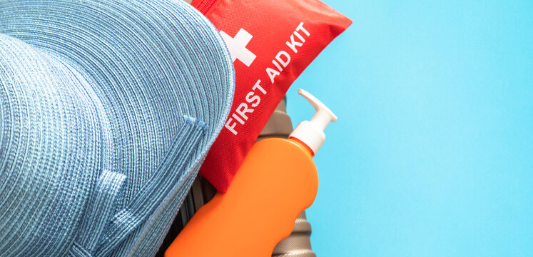 Red first aid kit, orange sunscreen bottle and straw hat on travel suitcase. Medical travel insurance concept, safe summer vacation close up banner with blue background and copy space for text.