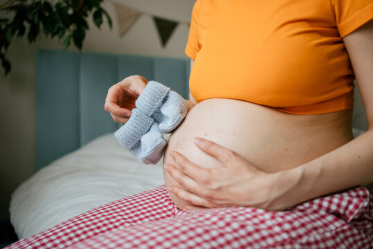 Pregnant woman anticipating motherhood, holding blue baby booties against her belly, preparing emotionally for the arrival of her future newborn child, feeling expectant love