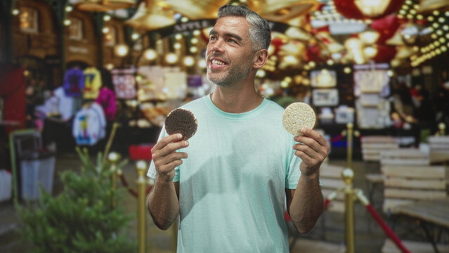Man holding two corn pastries in both hands with a cheerful smile amid festive market lights street; joyful choice.