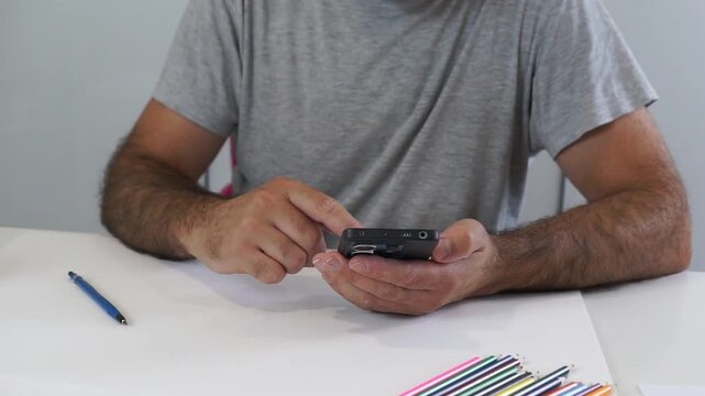 A man pauses his drawing activity to look at his smartphone, seeking reference images or taking a break next to colored pencils and paper.
