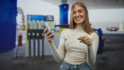 Young redhead woman smiling, holding smartphone, pointing finger at camera in gas station forecourt wearing light sweater and jeans  playful confidence. © Krakenimages.com